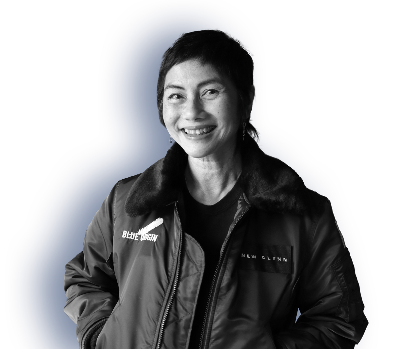 A woman with short hair smiles, standing with her hands in the pockets of a New Glenn branded jacket with a fuzzy collar.