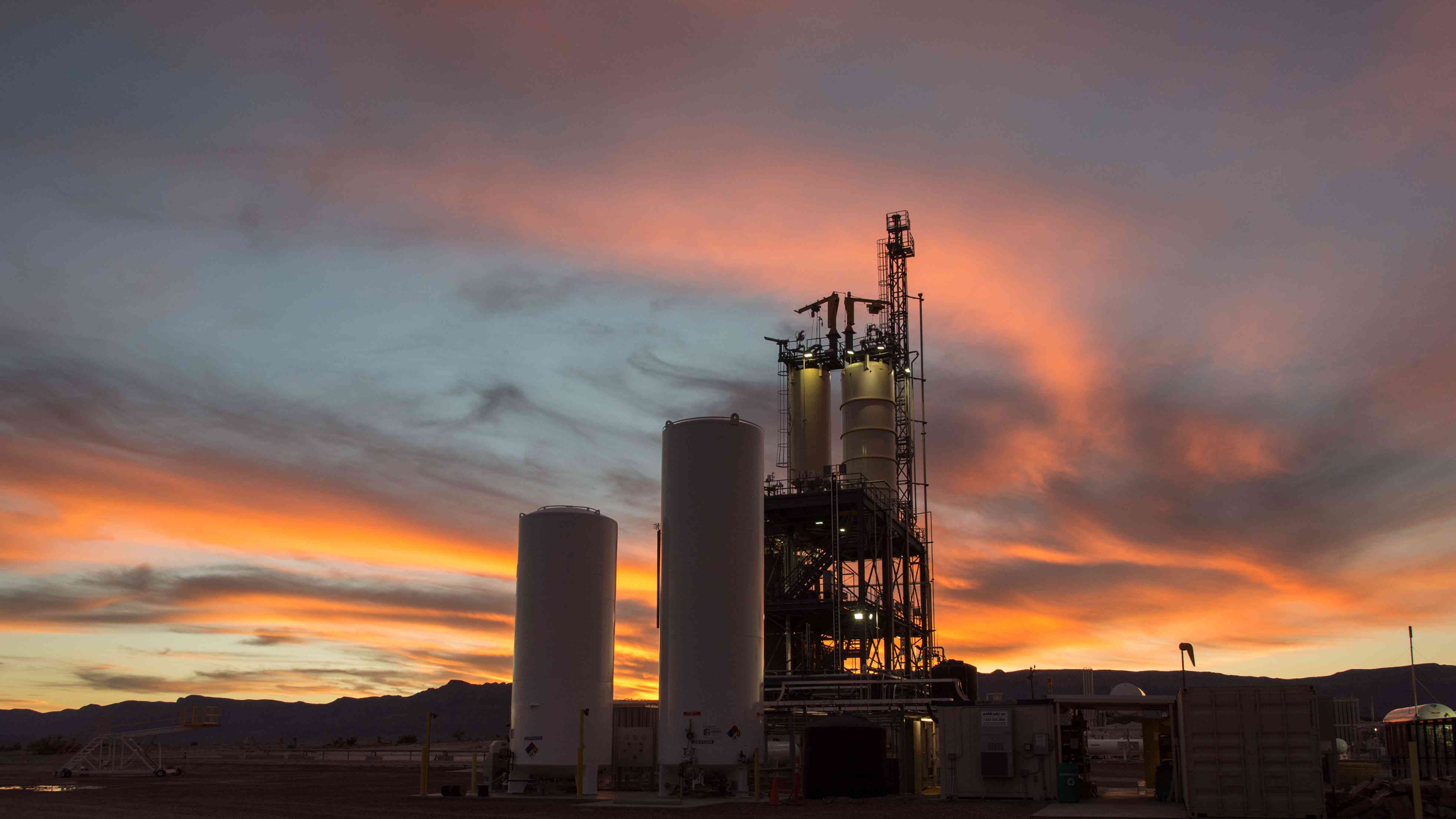 The towers of an engine test stand are visible in the distance against a bright pink and blue sky.