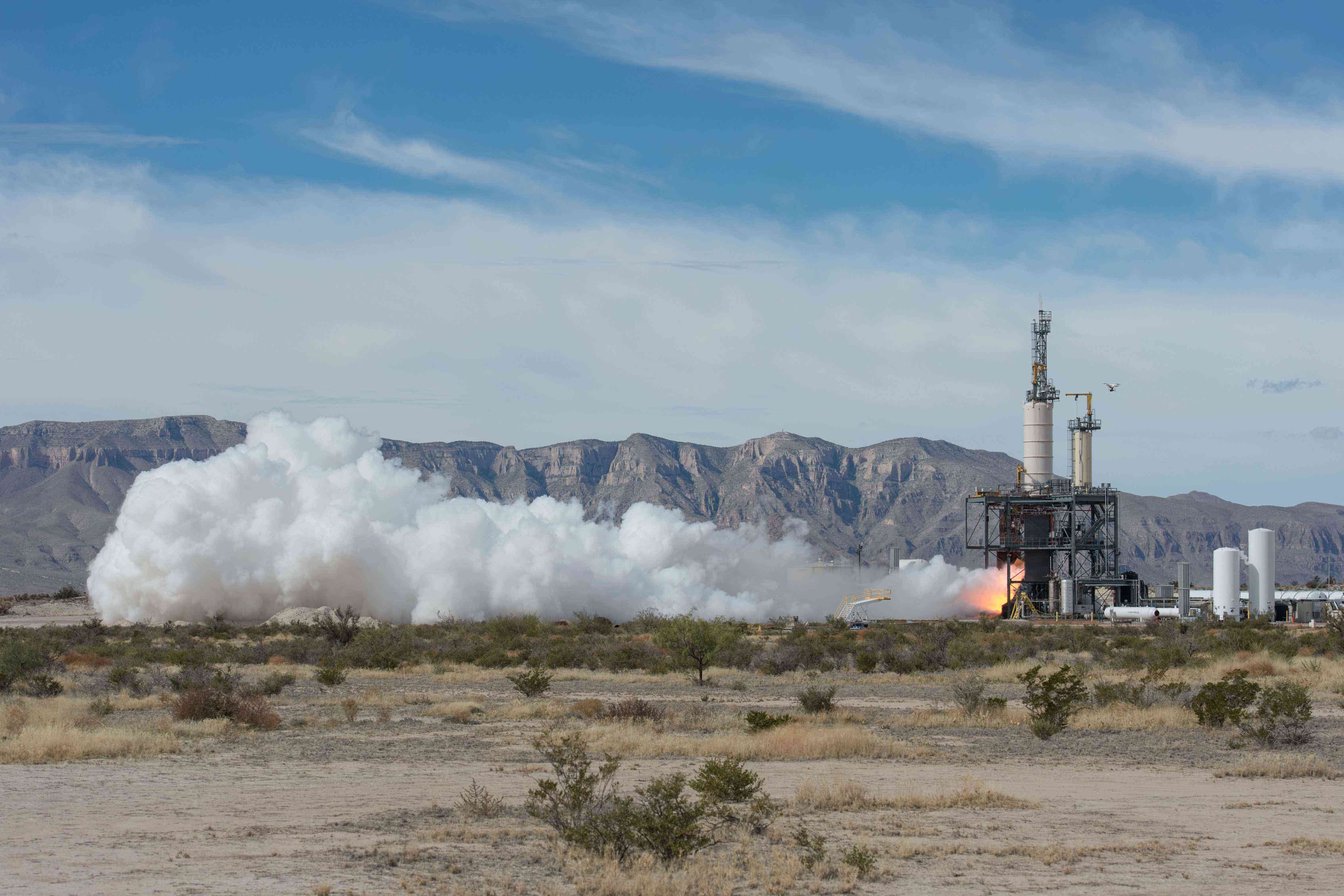 A BE-3 engine is hotfired on a test stand during the day, expelling a large cloud of horizontal dust and smoke seen from a distance.